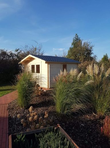 Klein Holzhaus umgeben von hohen Gräsern und Pflanzen unter blauem Himmel.